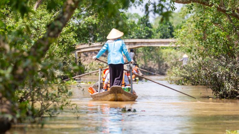 A traveler in a blue jacket and conical hat stands in a wooden boat navigating through dense mangrove forests in the Mekong