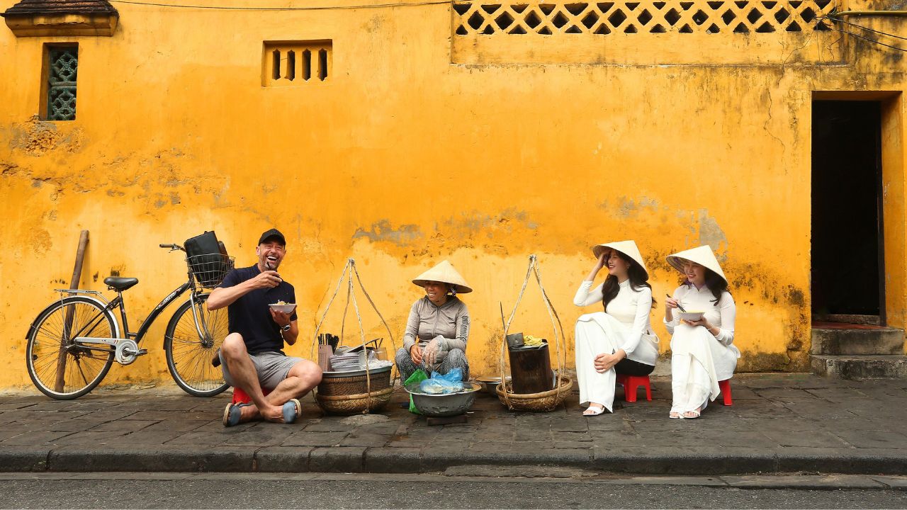 A cyclist interacting with local vendors wearing traditional hats in front of a bright yellow colonial building.