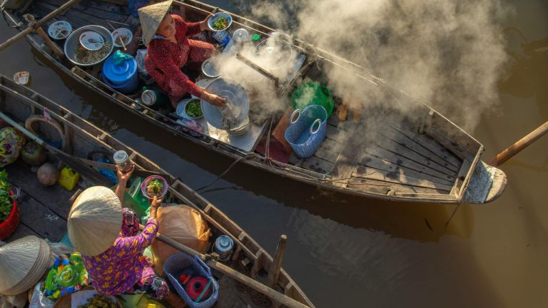 Overhead view of two wooden boats at a floating market with vendors cooking, steam rising, bowls and produce visible.