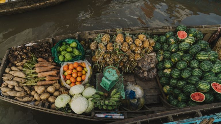 A wooden boat laden with fresh produce including pineapples, watermelons, cucumbers, and root vegetables at a floating