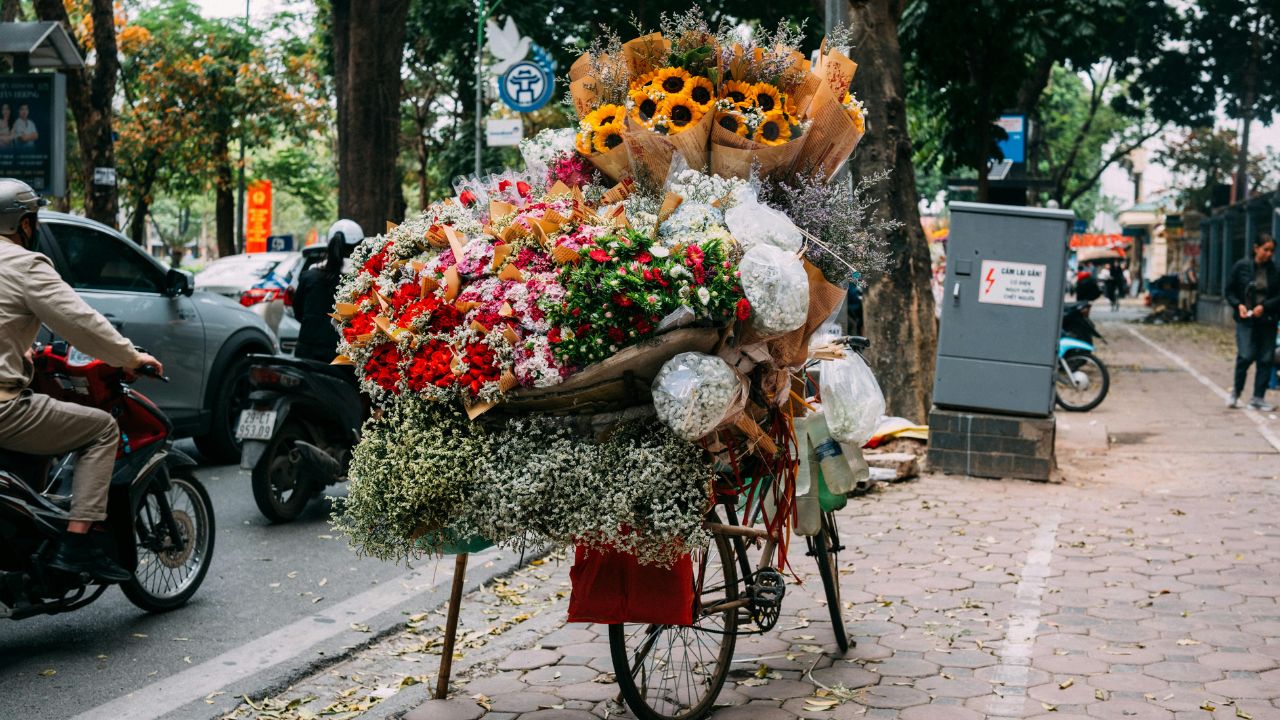 A flower vendor's bicycle loaded with colorful bouquets and sunflowers on a tree-lined Hanoi street.