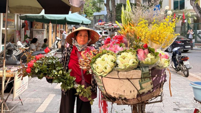 A local flower vendor in a red áo dài and conical hat selling fresh flowers from a bicycle cart on a Hanoi street.