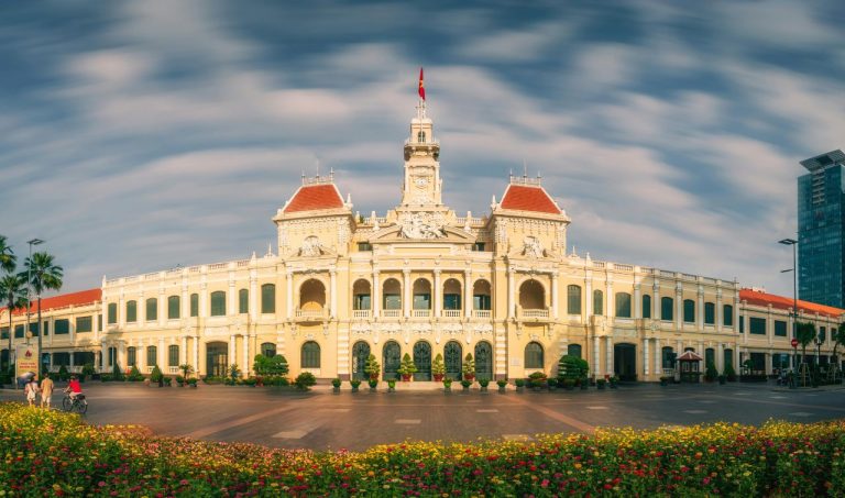 Yellow colonial city hall with red-roofed towers and central clock under blue sky, Ho Chi Minh City.