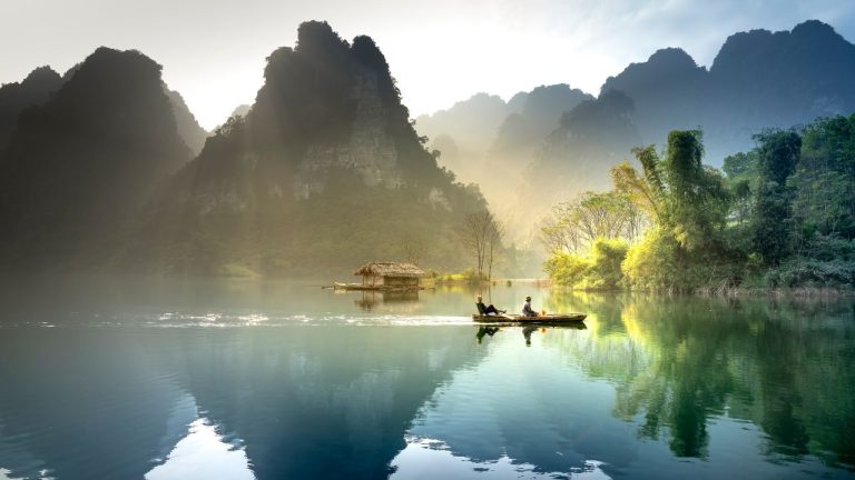 Two travelers paddling a boat on calm water at sunrise, with dramatic limestone karsts and mist in background.