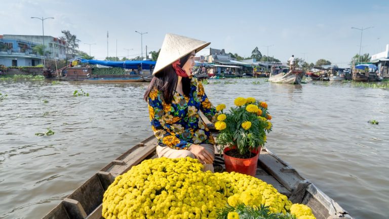 A vendor in a traditional conical hat selling yellow flowers from a wooden boat at a Mekong Delta floating market.