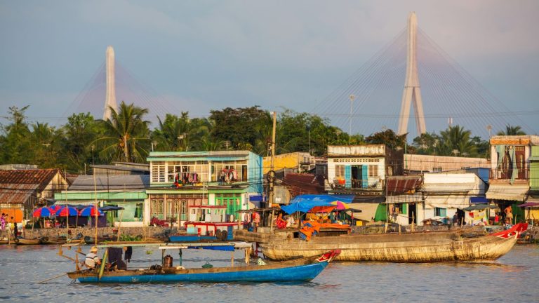 Riverside village with colorful wooden boats and houses along the Mekong Delta, with a cable-stayed bridge in the background.