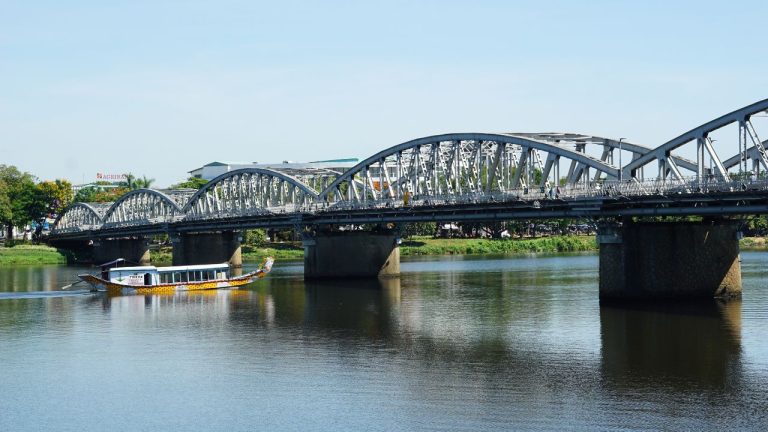 Metal arch bridge spanning a river with a traditional wooden boat passing underneath in Tours, Vietnam.
