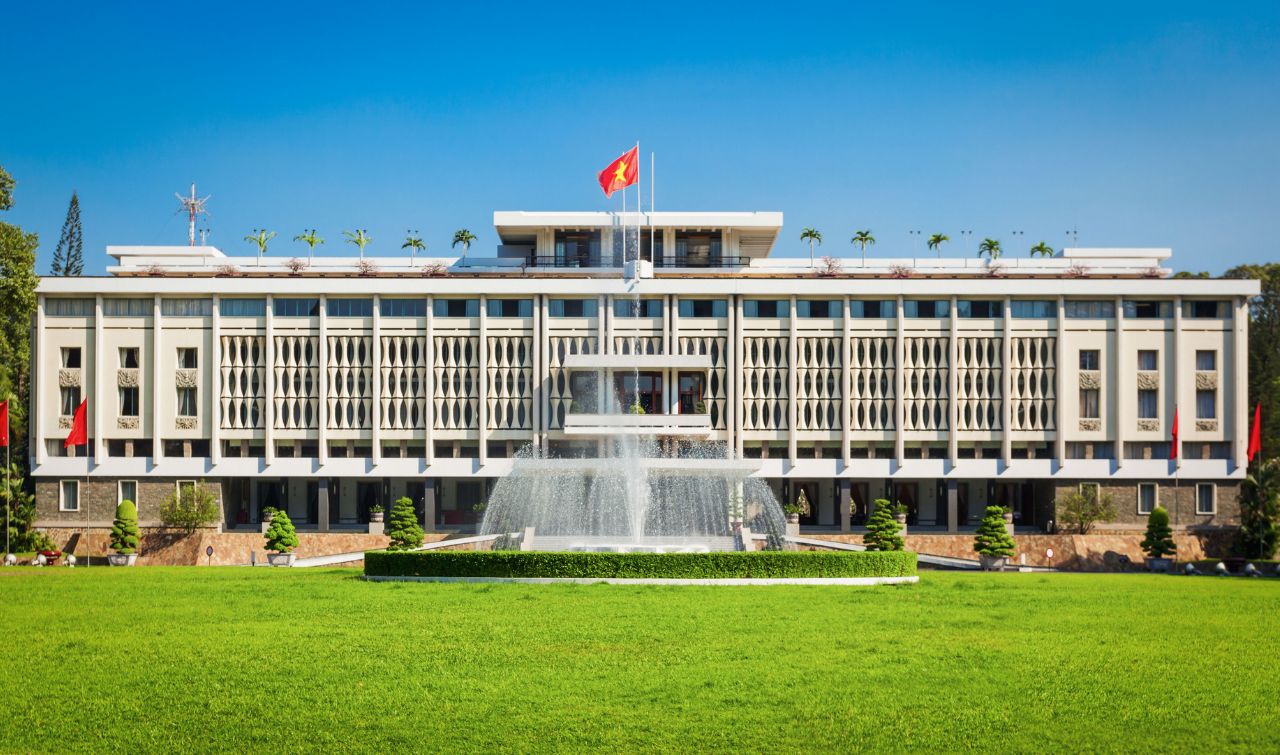 Reunification Palace with Vietnamese flag, fountain and manicured grounds under blue sky.