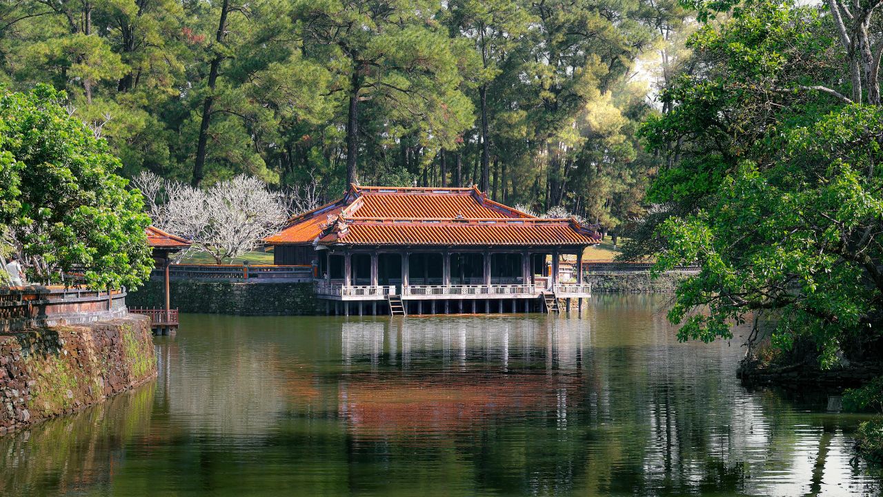 Traditional Vietnamese temple with orange tile roof reflected in calm water, surrounded by lush green forest.