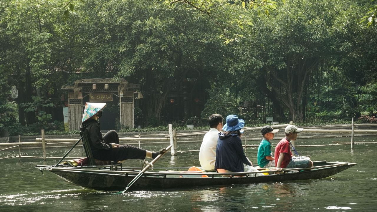 Travelers in a traditional wooden boat on calm water beneath lush trees in Ninh Binh.