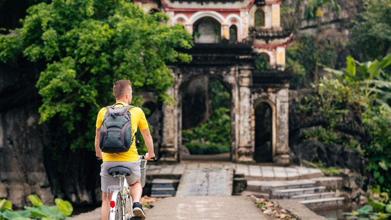 A cyclist in a yellow shirt rides toward an ivy-covered ancient temple with red-tiled roof in Ninh Binh, Vietnam.