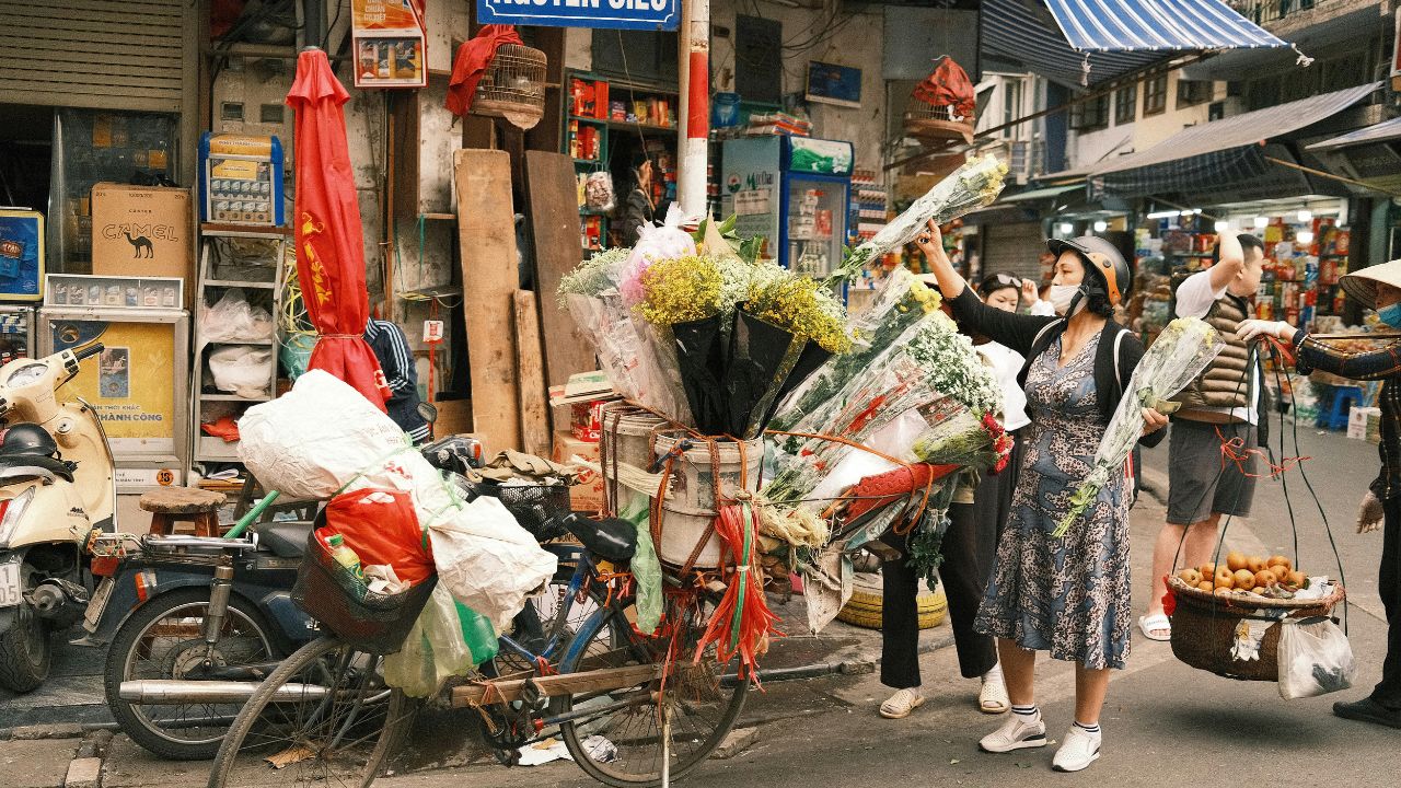 A flower vendor arranges colorful bouquets on a bicycle in a bustling Hanoi street market.