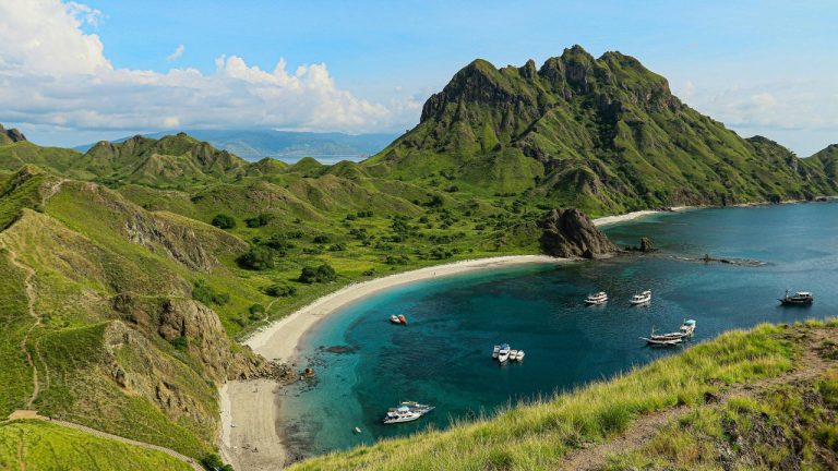 Dramatic green mountainous islands with sandy beach cove and turquoise water dotted with fishing boats in Halong Bay.