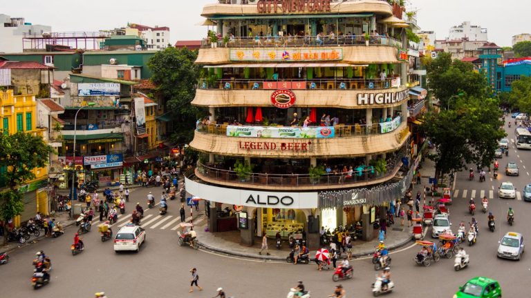 Busy street intersection in central Hanoi with circular multi-story building and scooter traffic.
