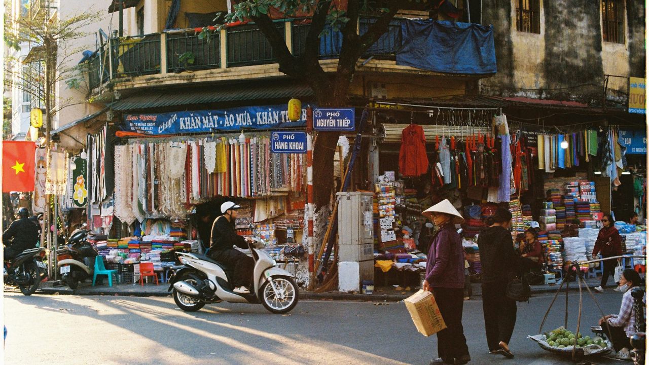 A busy street market in Hanoi with vendors, shops displaying textiles and goods, a scooter passing by, and shoppers browsing.