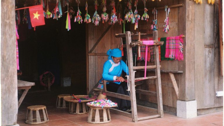 Local artisan in blue clothing working with colorful woven textiles and traditional crafts displayed in a wooden workshop.