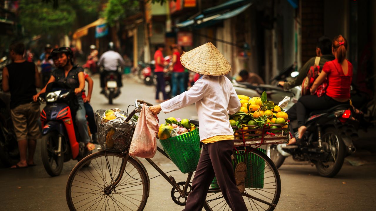 A street vendor in a conical hat selling yellow fruit from a bicycle on a busy Hanoi street.