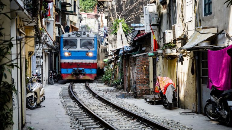 A blue and red train moving through a narrow residential alley lined with old buildings and scooters in Hanoi.
